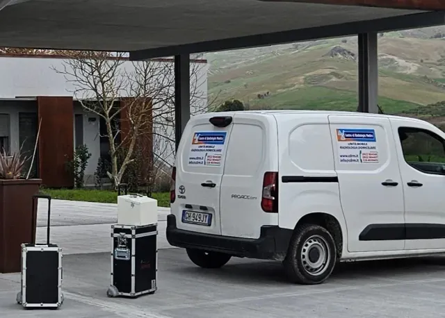 Un furgone bianco di servizio è parcheggiato accanto alle pompe di benzina, sotto una tettoia, con le colline sullo sfondo.