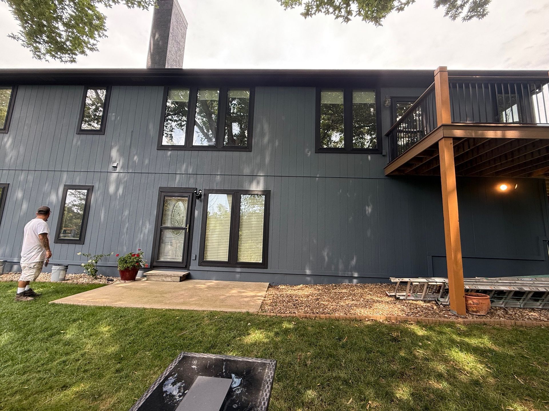 Person painting dark blue exterior wall of a two-story house with black trim, deck, and chimney.