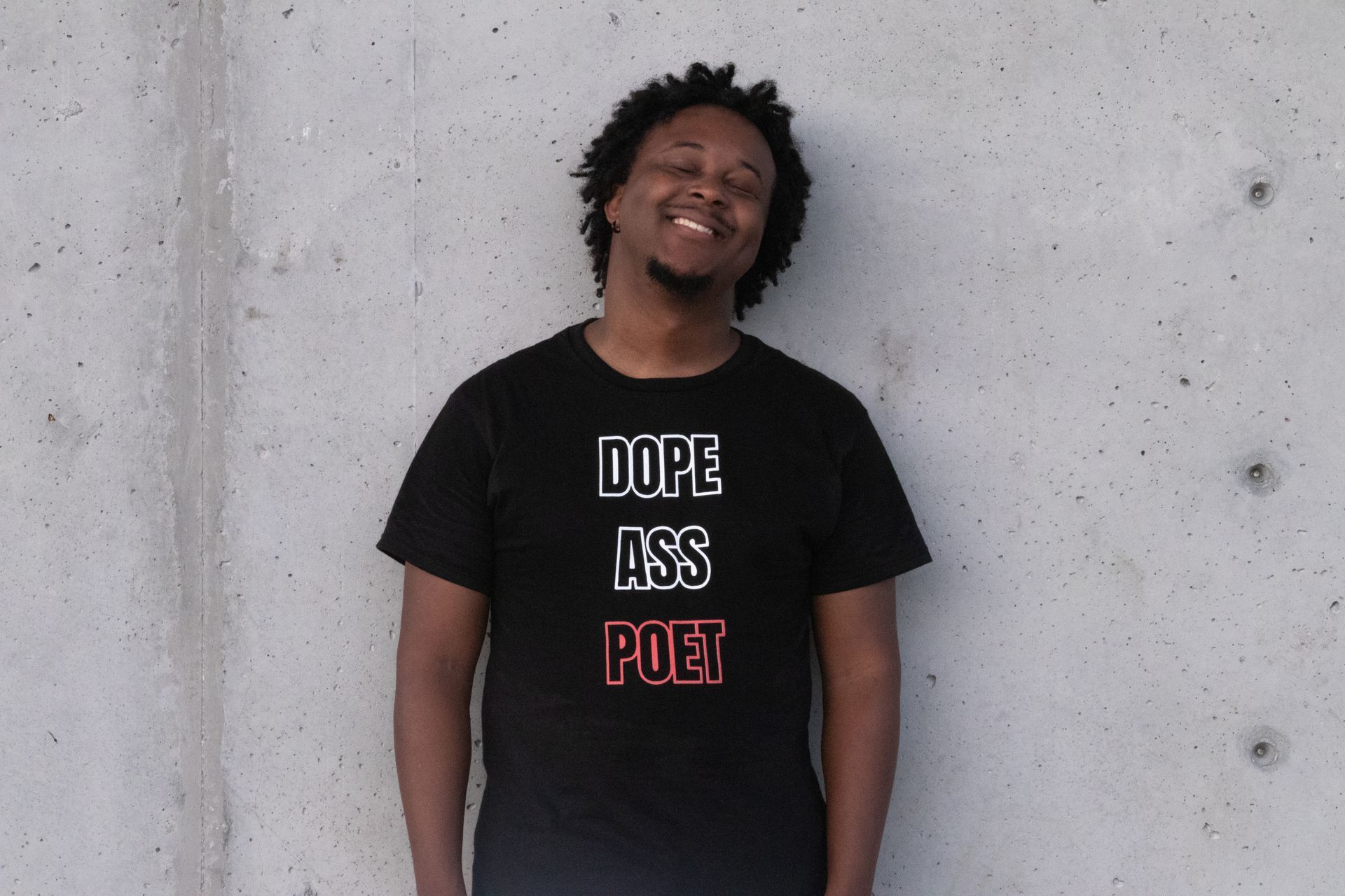 A person standing in front of a concrete wall smiling while wearing a t-shirt that says