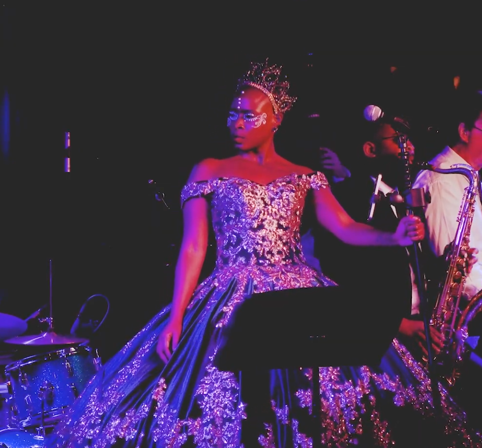 Woman in gown with band, singing at a concert with a mask. Stage lit with purple and blue lights.