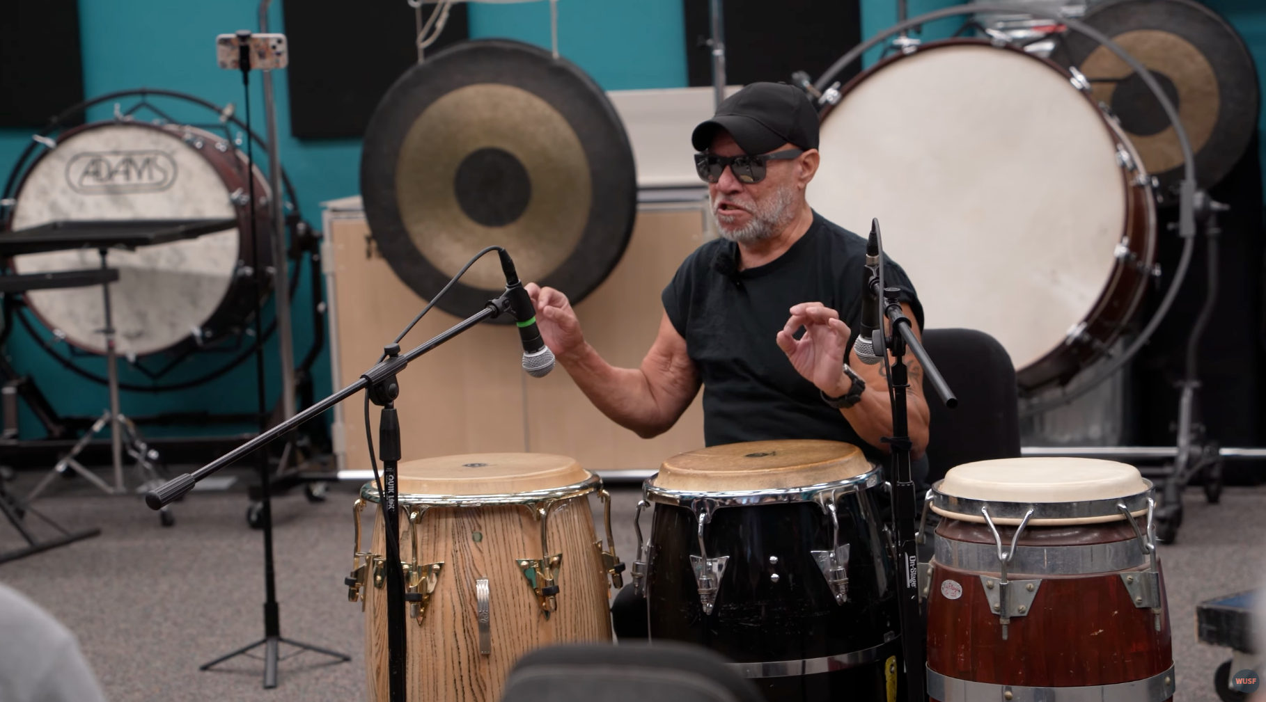Man in a black shirt gestures, seated by conga drums. Percussion instruments surround him in a studio.