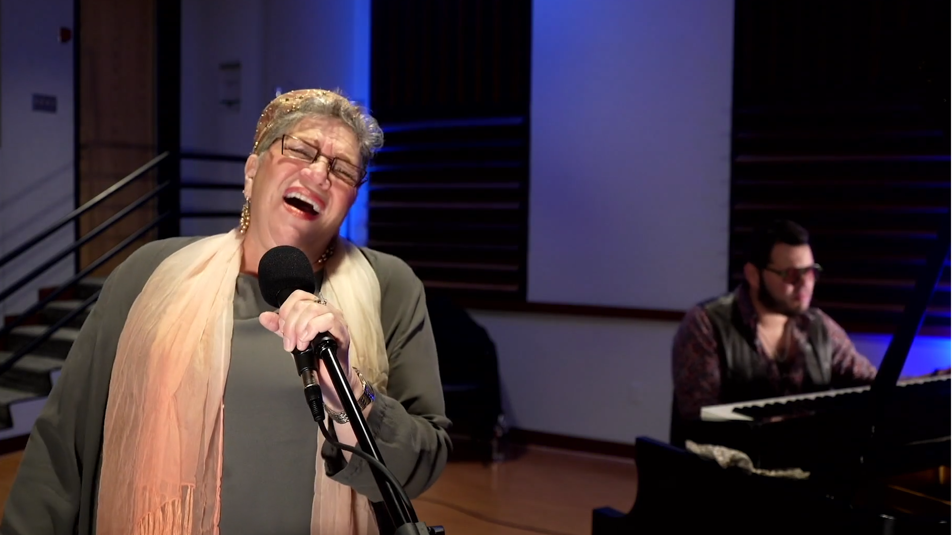 A woman is singing into a microphone in front of a man playing a piano.