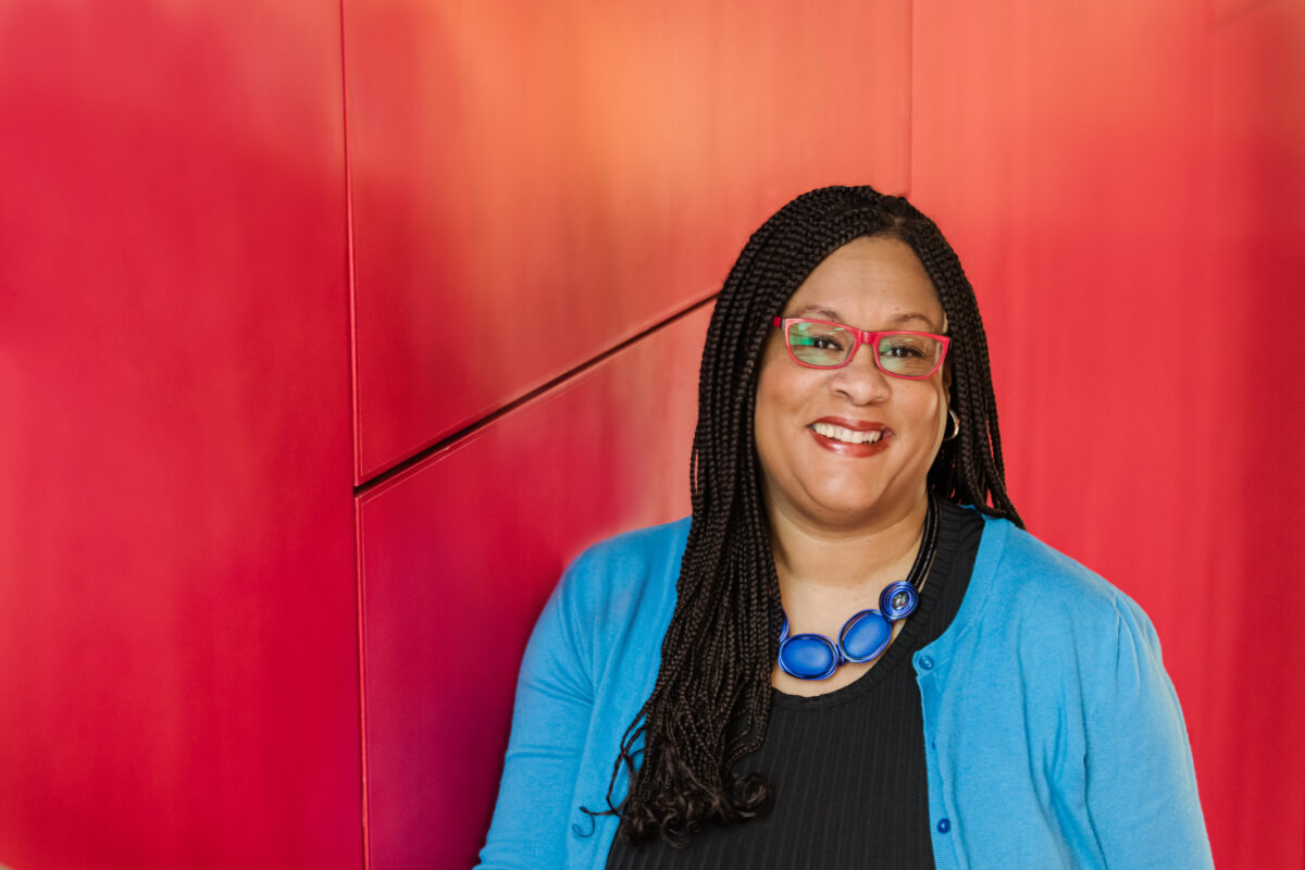 A woman wearing glasses and a blue sweater is smiling in front of a red wall.