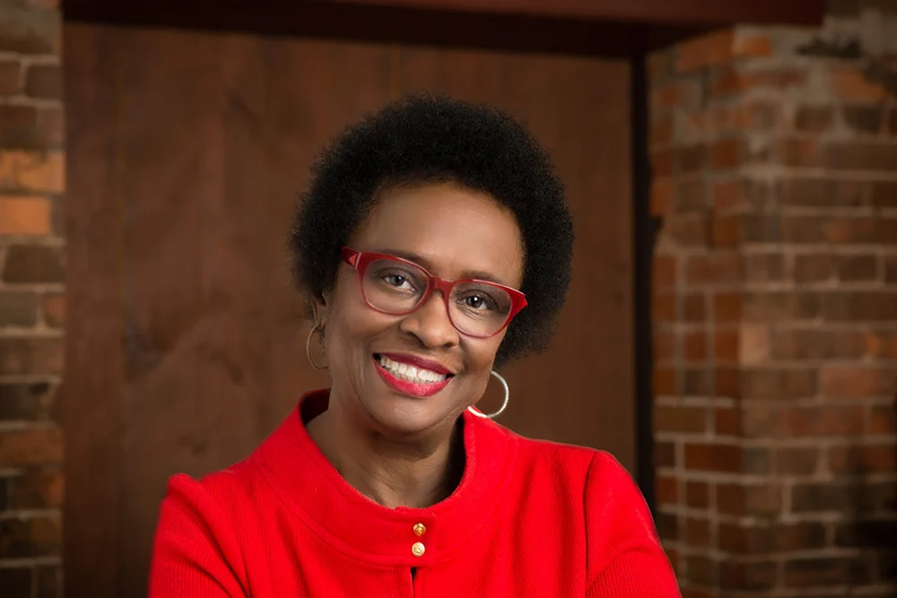 A woman wearing glasses and a red shirt is smiling in front of a brick wall.