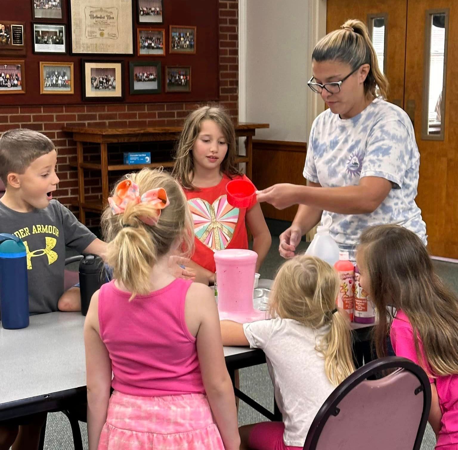 A group of children are sitting around a table with a woman in a tie dye shirt