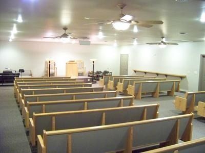 Empty church sanctuary with rows of wooden pews, ceiling fans, and a small altar at the front.