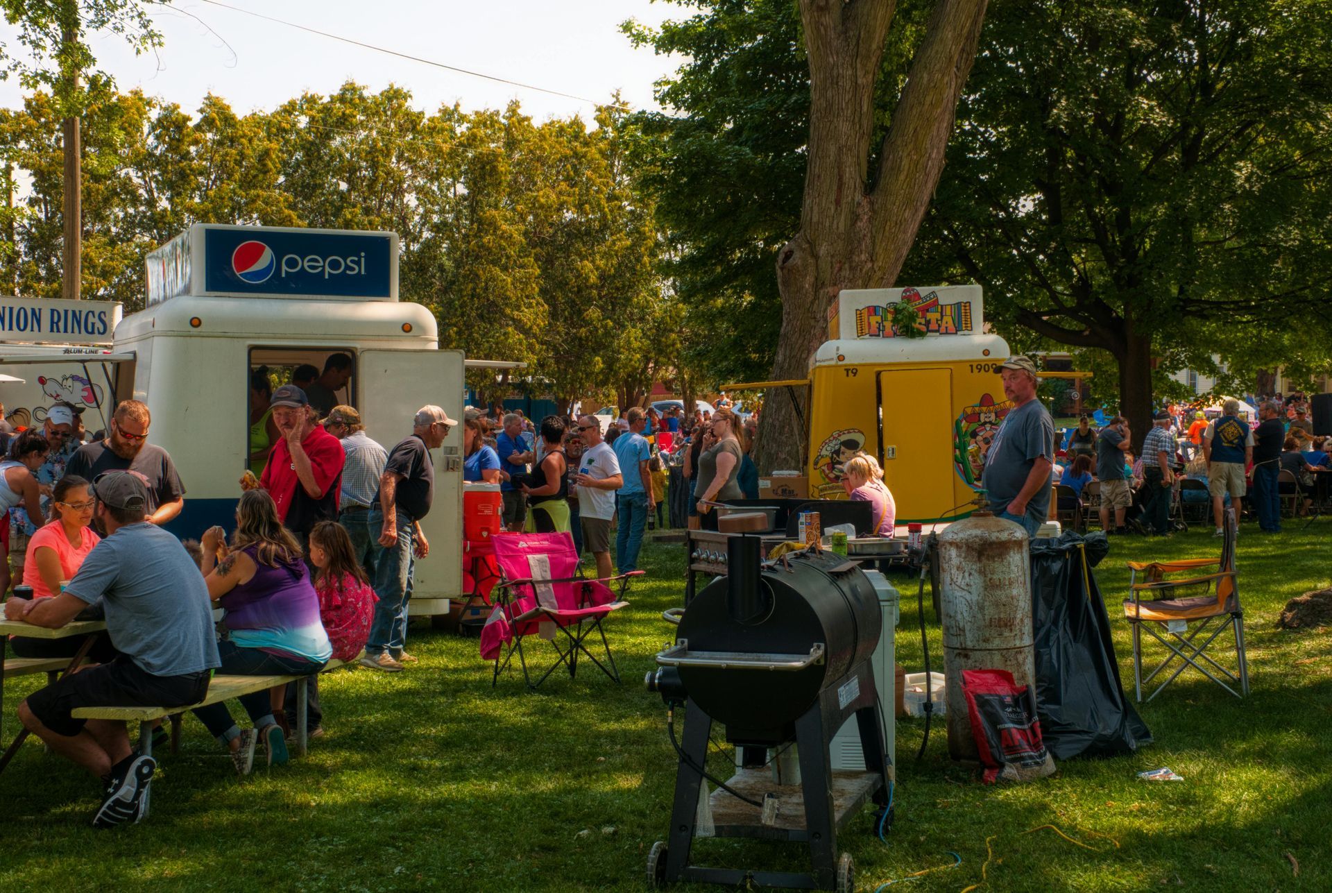 People at an outdoor park event with food stalls, tables, and trees in warm sunlight