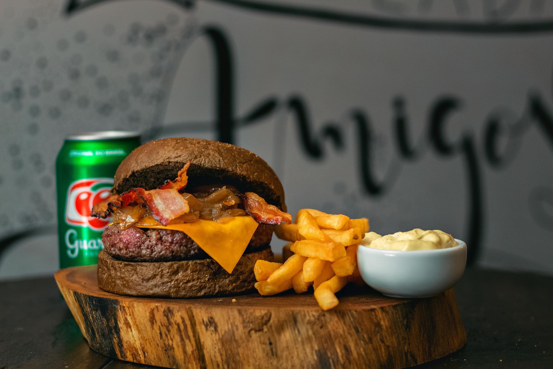 Burger with cheese, bacon, fries, and a side of sauce on a wooden board with a soda can behind it