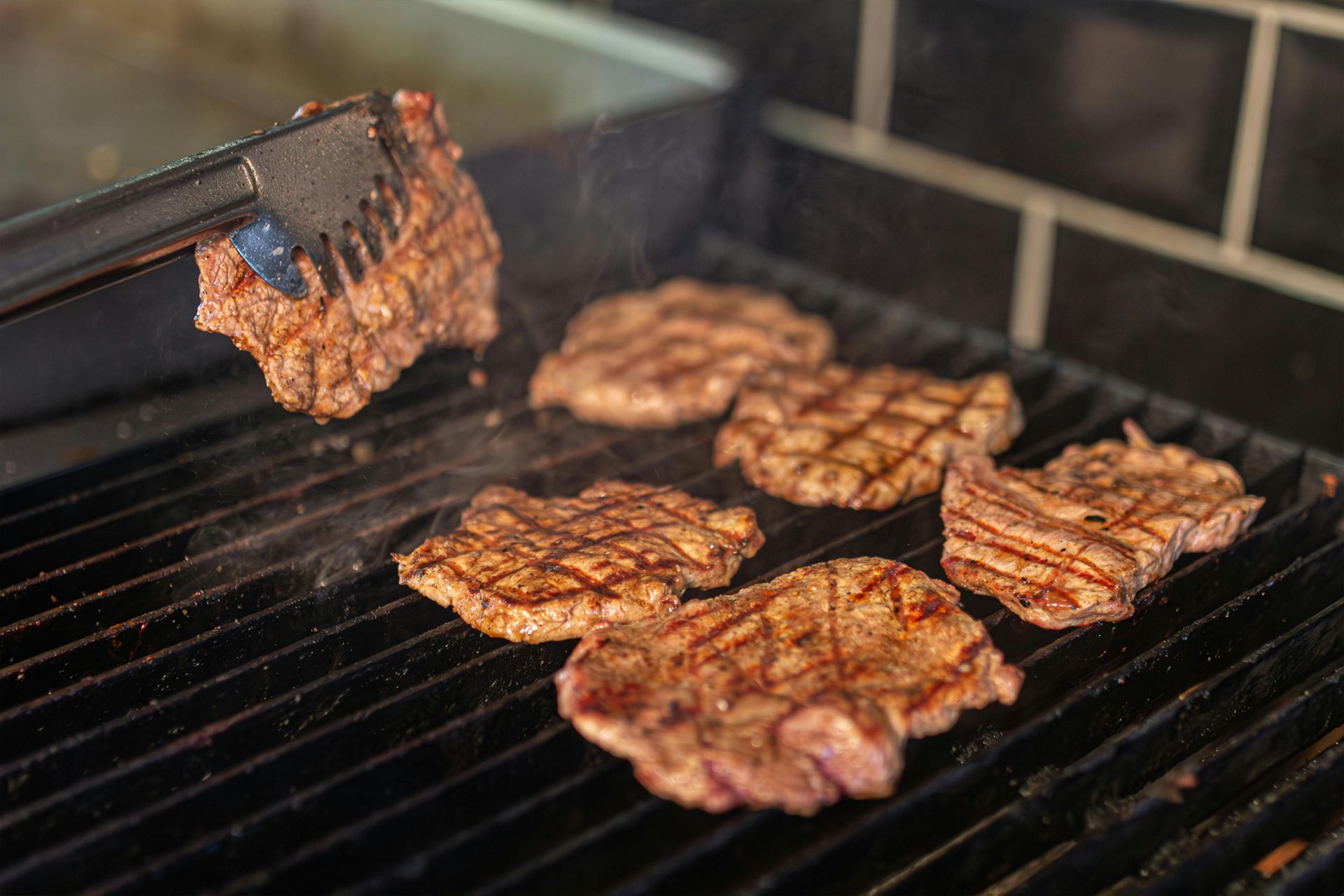 Steaks sizzling on a grill, with one piece being flipped by tongs.
