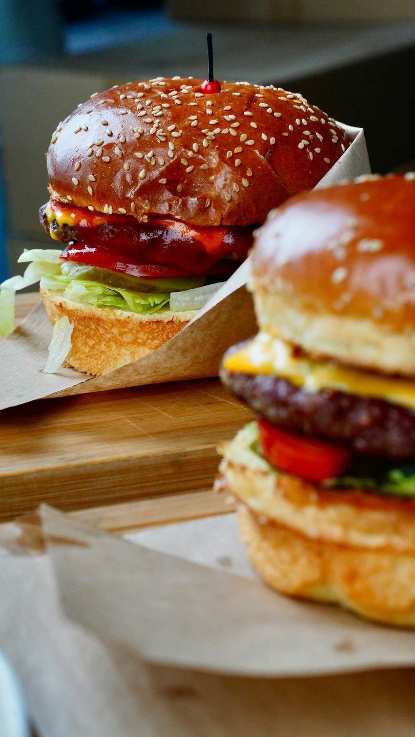 Two cheeseburgers with sesame buns on parchment paper, one in focus on a wooden board.