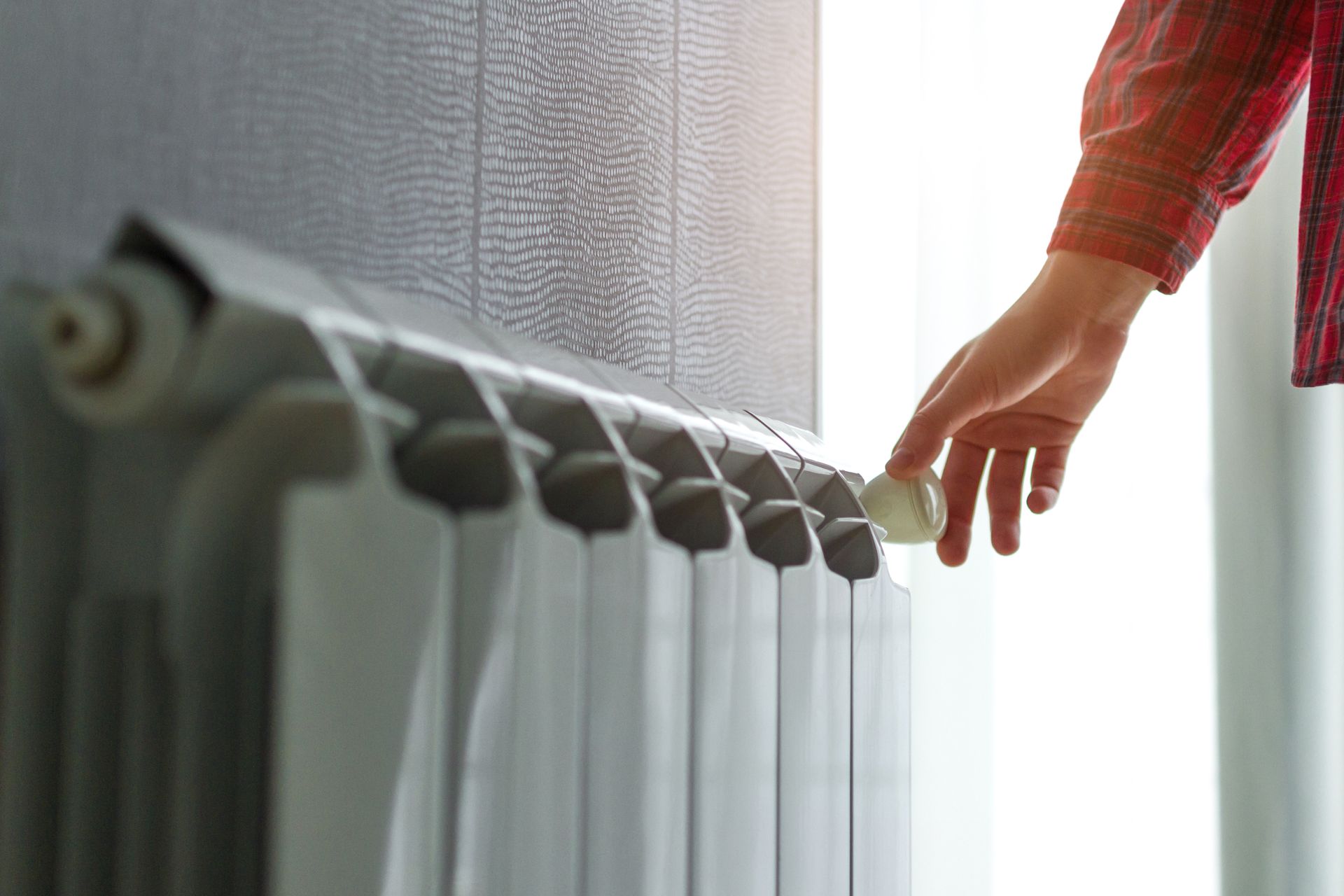 Main d'une personne réglant un robinet de radiateur, près d'une fenêtre. Chemise à carreaux rouges. Couleurs blanc et gris.