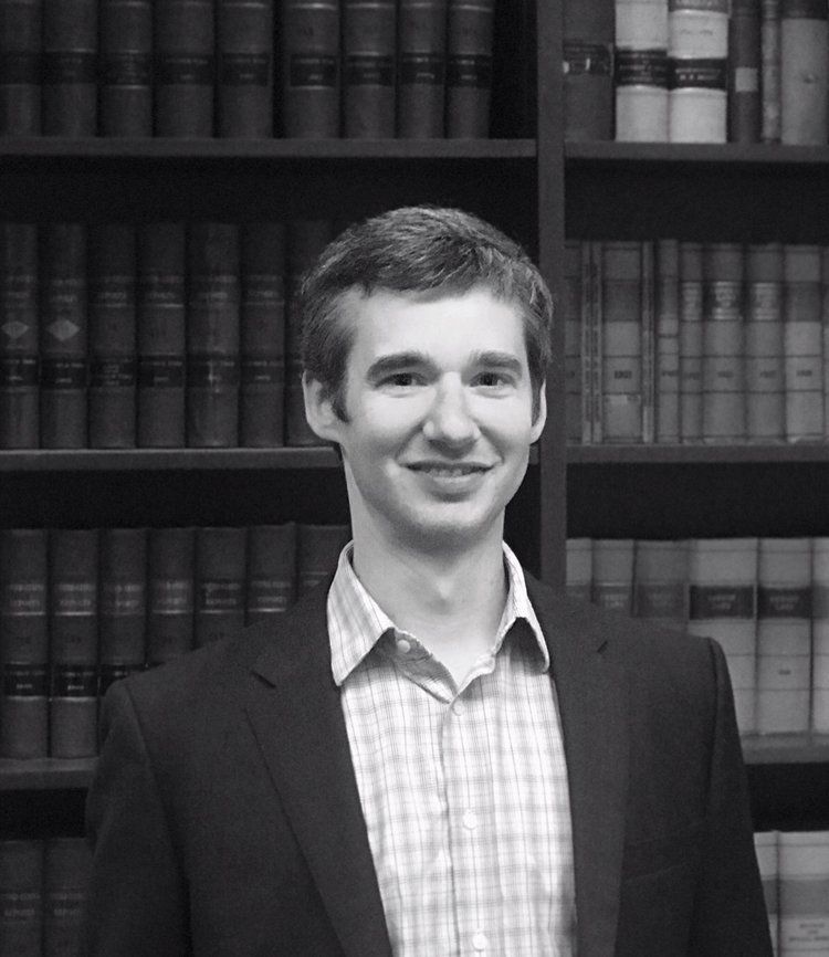 a man in a suit and plaid shirt stands in front of a bookshelf