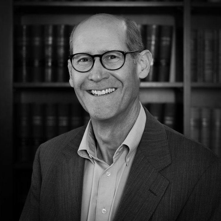 a man wearing glasses and a suit smiles in front of a bookshelf