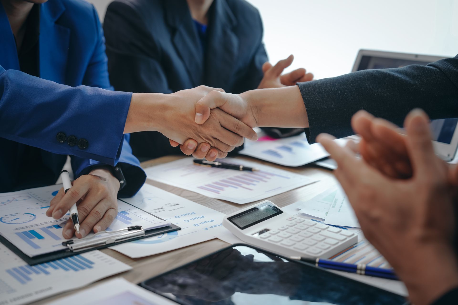 Business handshake with documents, calculator, and laptop on table.