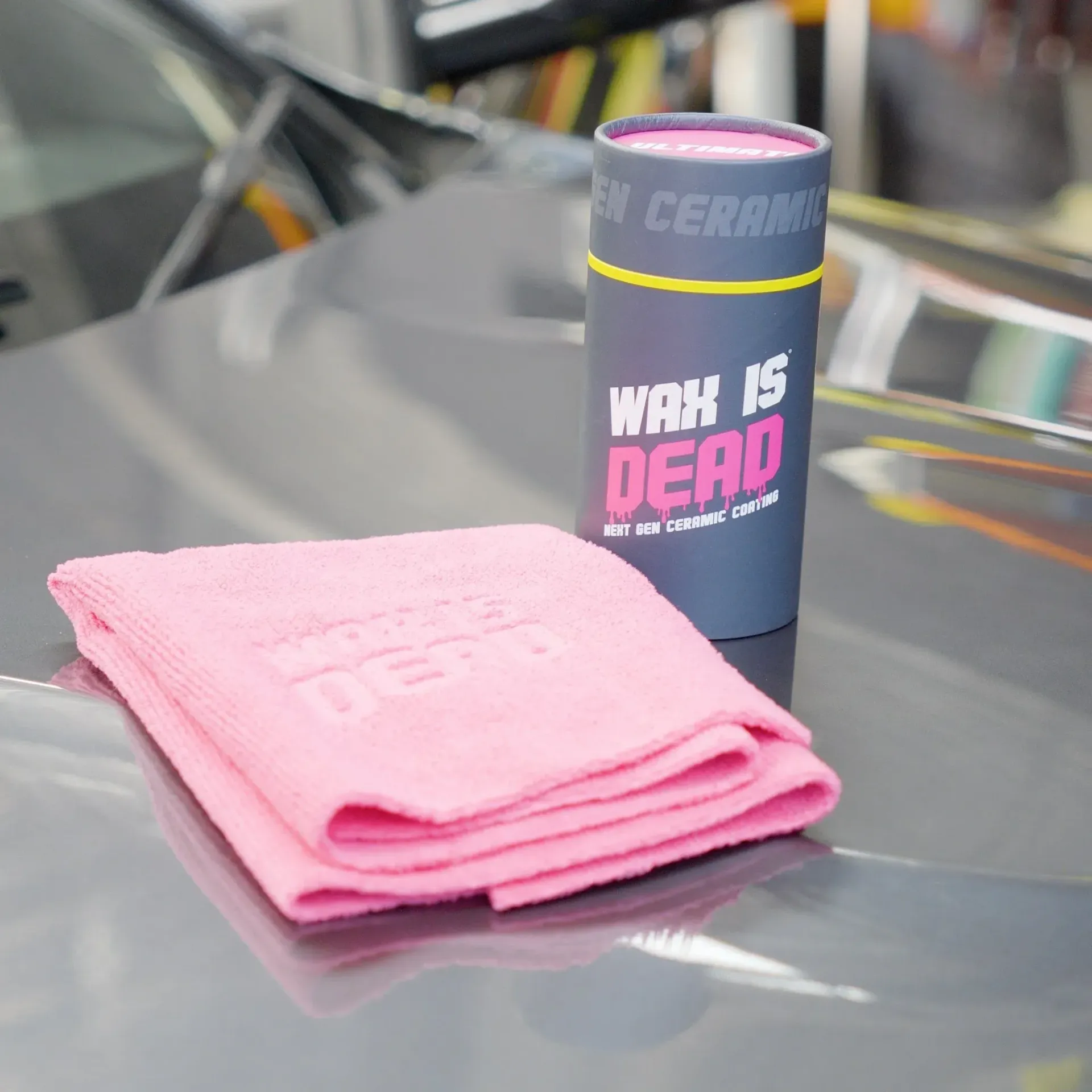 Pink microfiber cloth beside a black ceramic mug on a car hood, with “WAX ON, WAX OFF” text on the mug