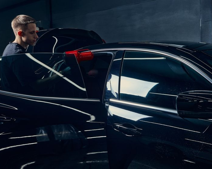 Person polishing a black car in a dim garage, with red taillights reflecting on the body.