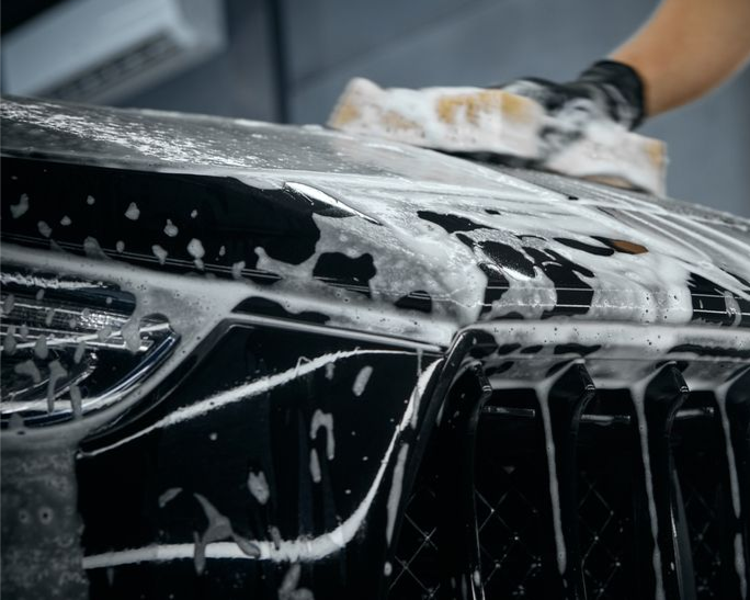 Black car front being washed with soapy foam by a gloved hand