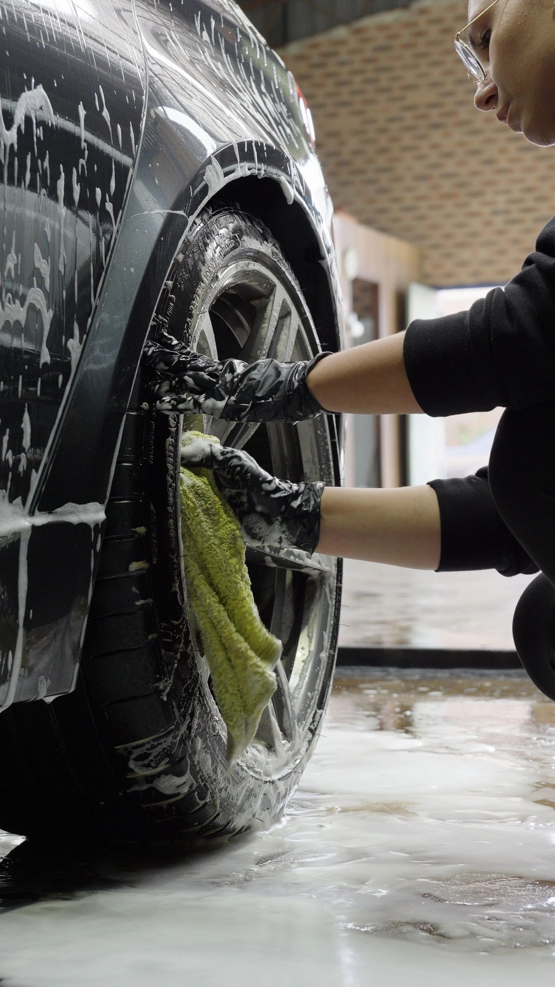 Person washing a black car wheel with a green cloth in a soapy garage.