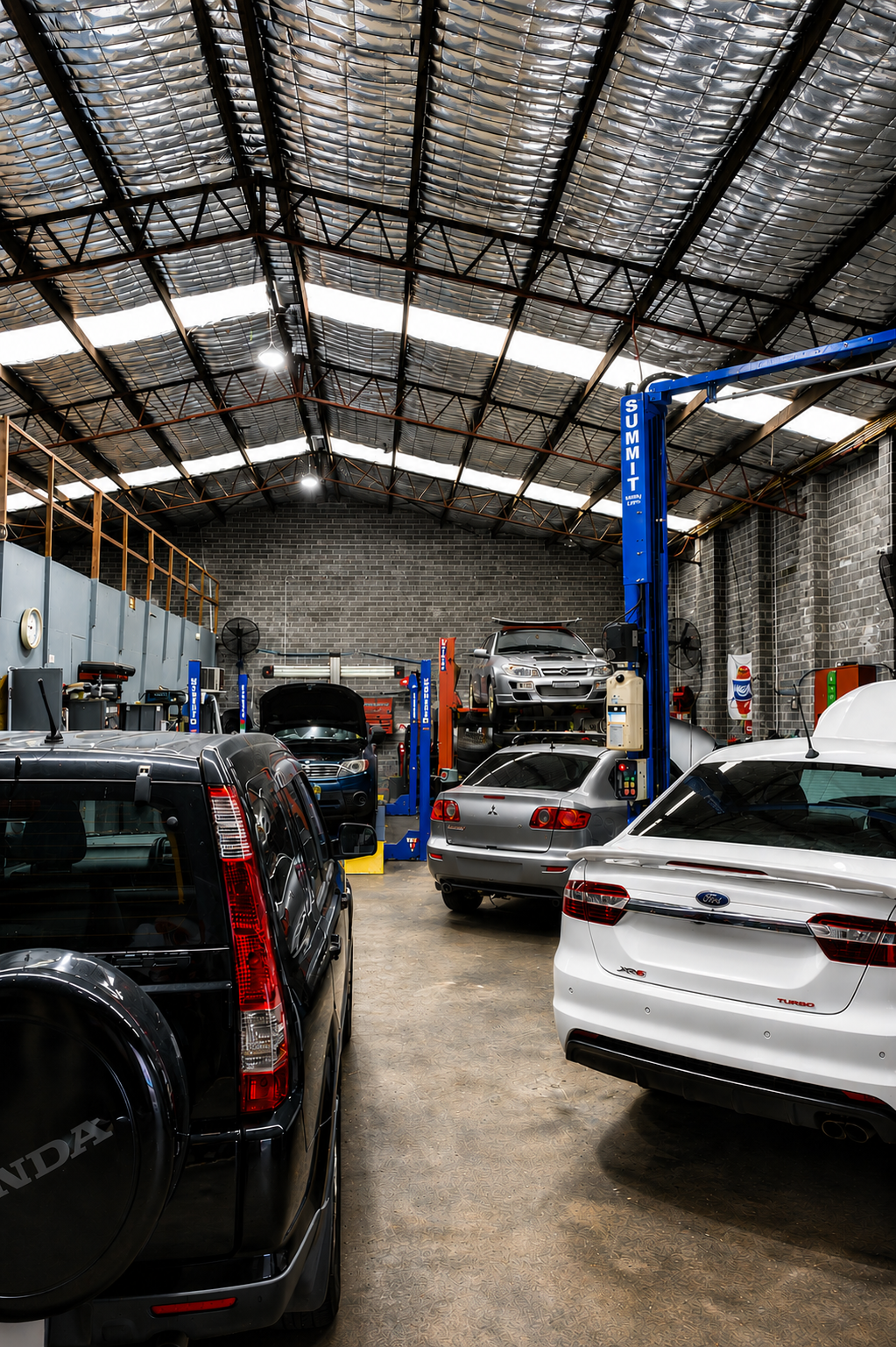Automotive repair shop interior with several cars parked under a high metal roof, including a blue lift and a white SUV