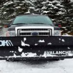 A black truck with a snow plow on the front is parked in the snow.