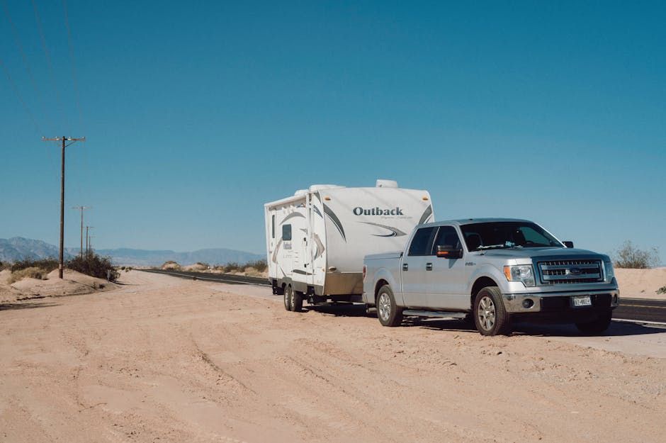 A silver truck is towing a trailer down a dirt road.