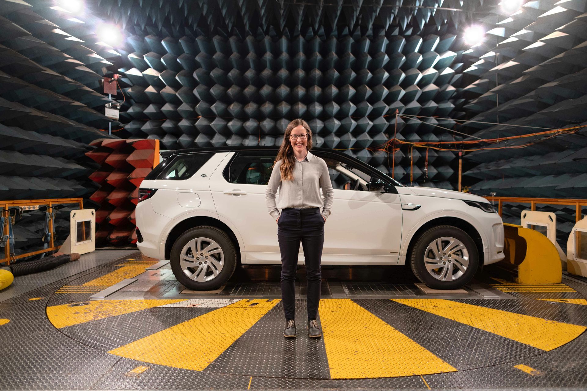 A woman is standing in front of a car in an acoustic room.