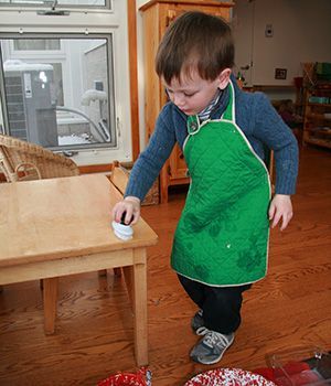 A young boy wearing a green apron is cleaning a wooden table.