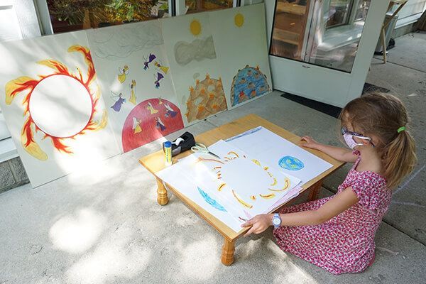 A little girl wearing a mask is sitting at a table drawing on a piece of paper.