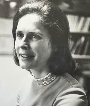 A black and white photo of a woman smiling in front of a bookshelf.