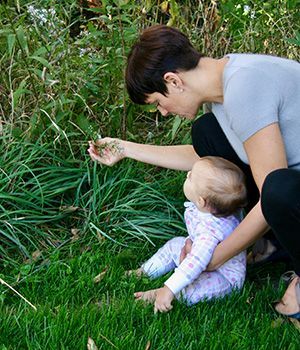 A woman kneeling on grass with a baby in arms shwing her some leaves