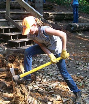An adolescent girl wearing a cap is cutting wood with a big axe.