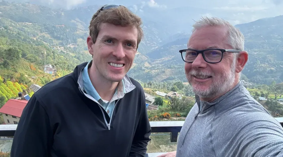 Two men are standing next to each other in front of a mountain.