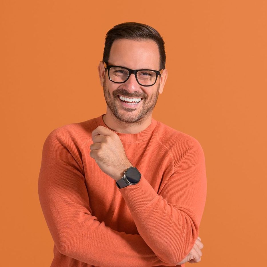 A man wearing glasses and an orange sweater is smiling with his arms crossed.