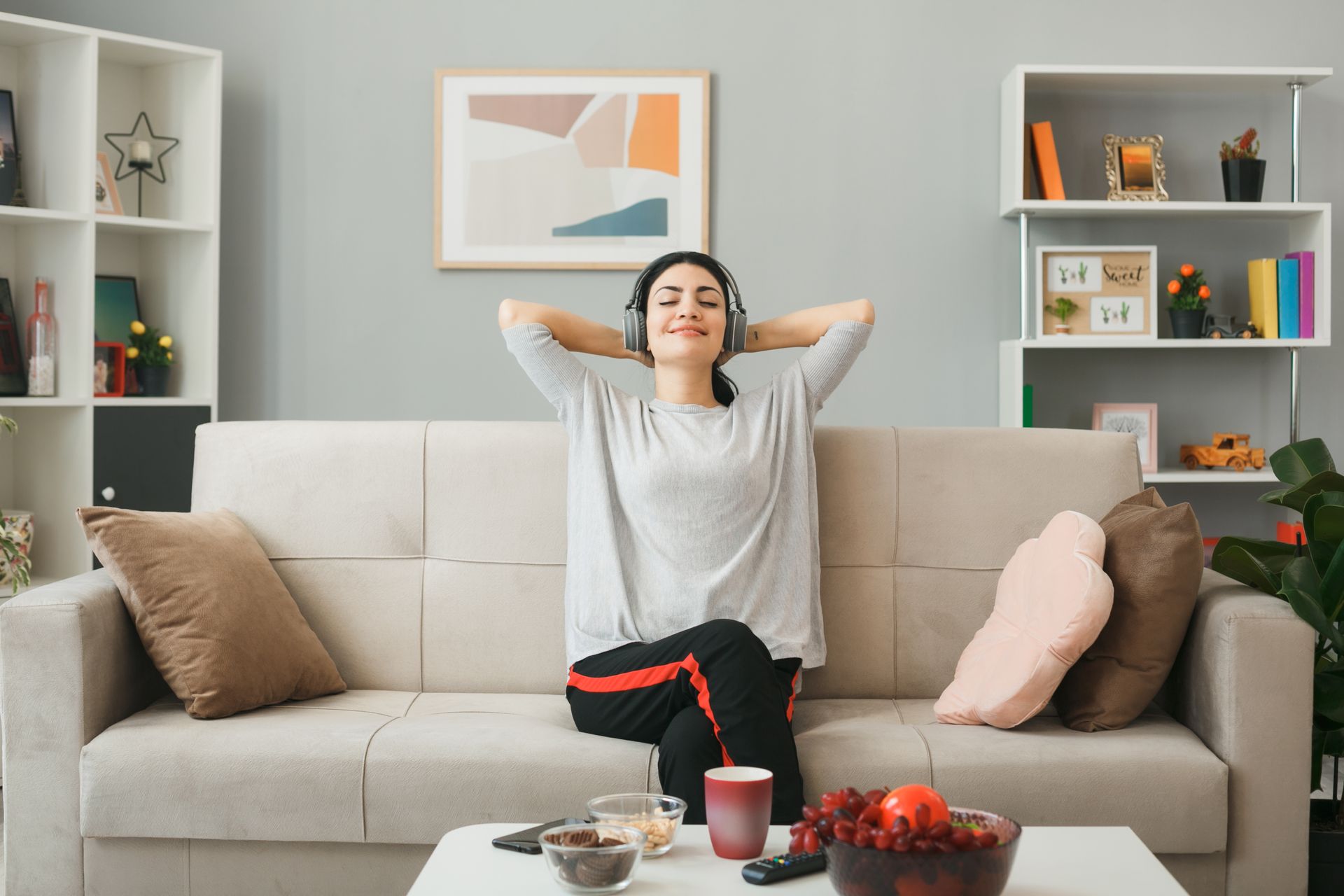 Person relaxing on a couch with hands behind head, in a bright living room with shelves and a coffee table.