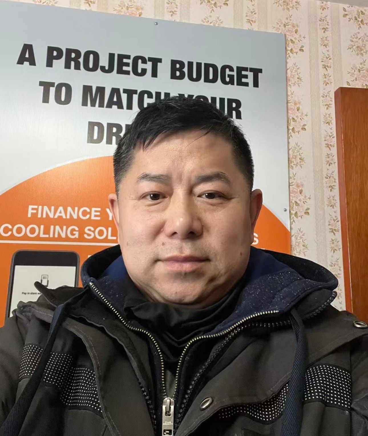 Man in a black jacket posing indoors in front of a sign with bold text and an orange circle.