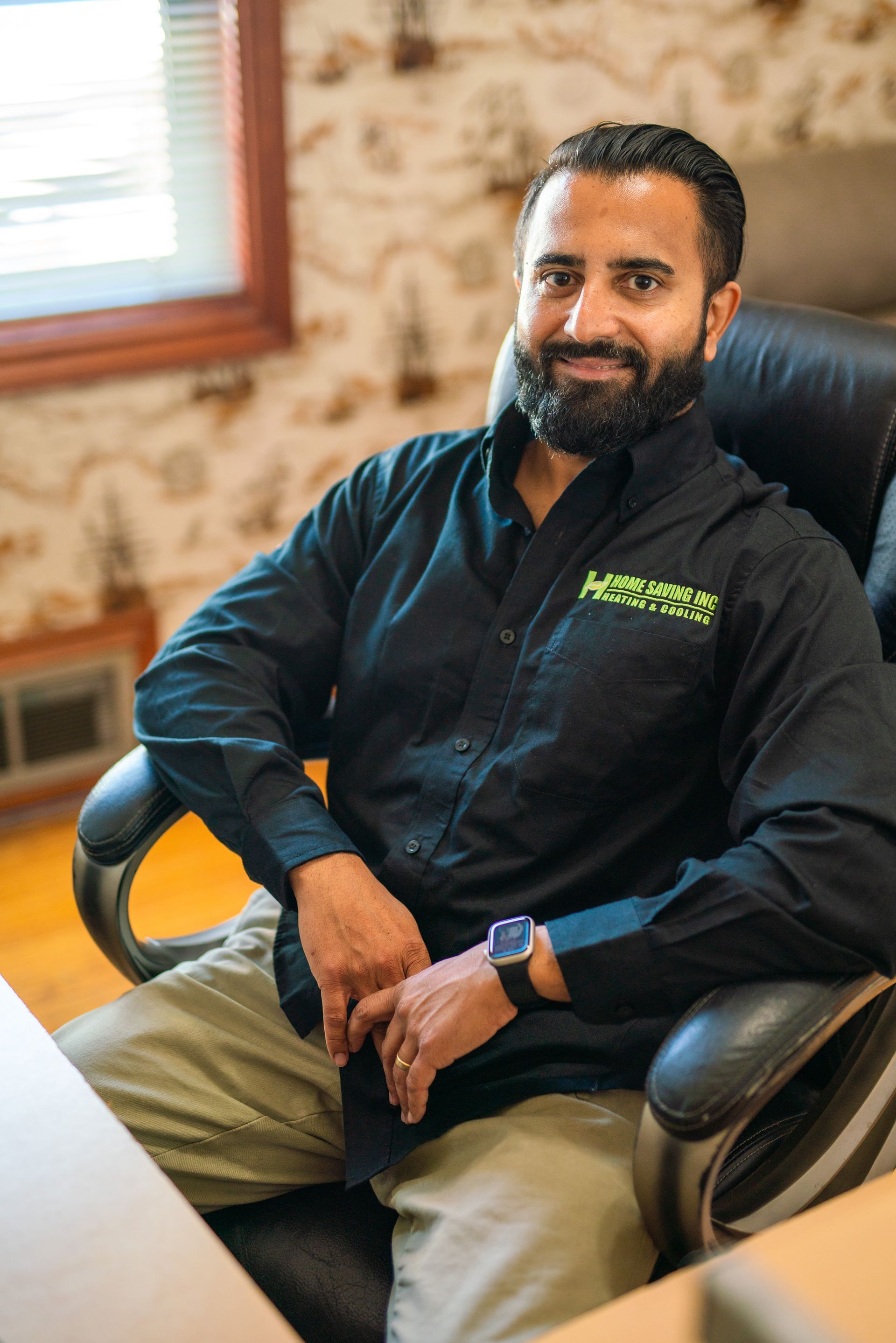 Man seated in an office chair, wearing a black shirt and khaki pants, smiling at a desk.