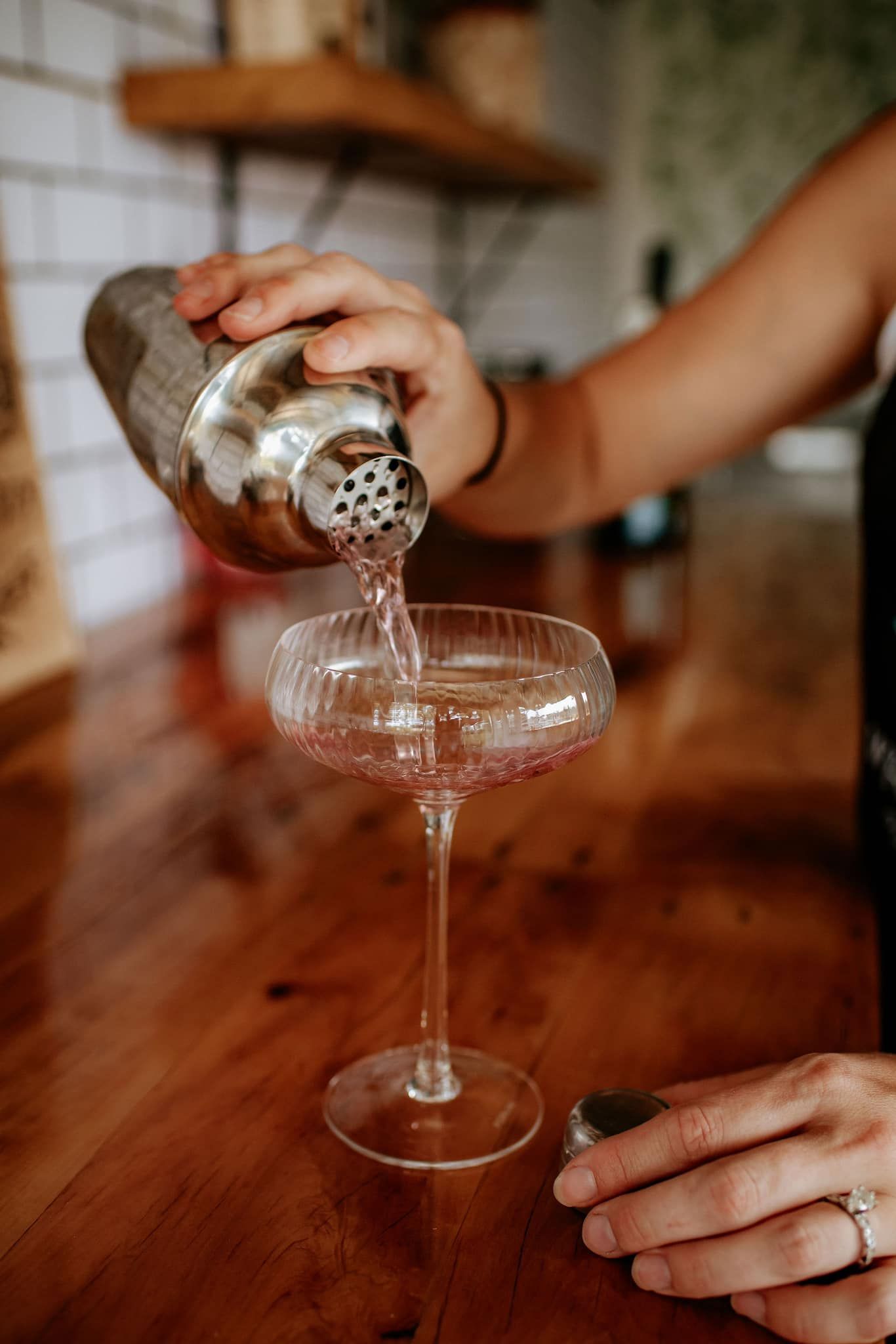 Person pouring a pink cocktail from a shaker into a stemmed glass on a wood surface.