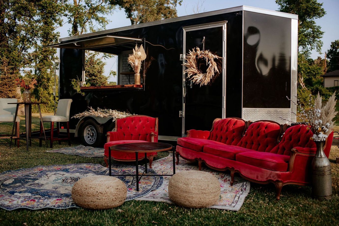 Outdoor lounge setup with red velvet sofa, chairs, rugs, and a black trailer decorated with wreaths.