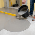 Gray floor leveling compound being poured from a black bucket onto a concrete floor.