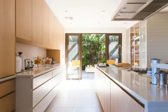 Bright modern kitchen with light wood cabinets, white countertops, and open doors to a garden patio.