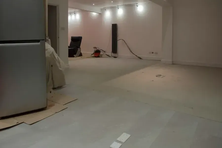 Empty room under renovation with exposed flooring, ceiling lights, and a refrigerator on the left
