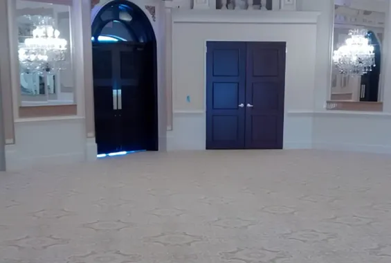 Empty elegant room with carpet, arched doorway, dark double doors, and chandeliers on both sides.