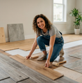 Person installing wood flooring in a bright room, kneeling and smiling with tools nearby