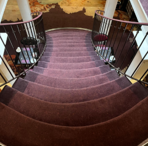 Curved carpeted staircase with dark railings and white columns in a warm-lit interior