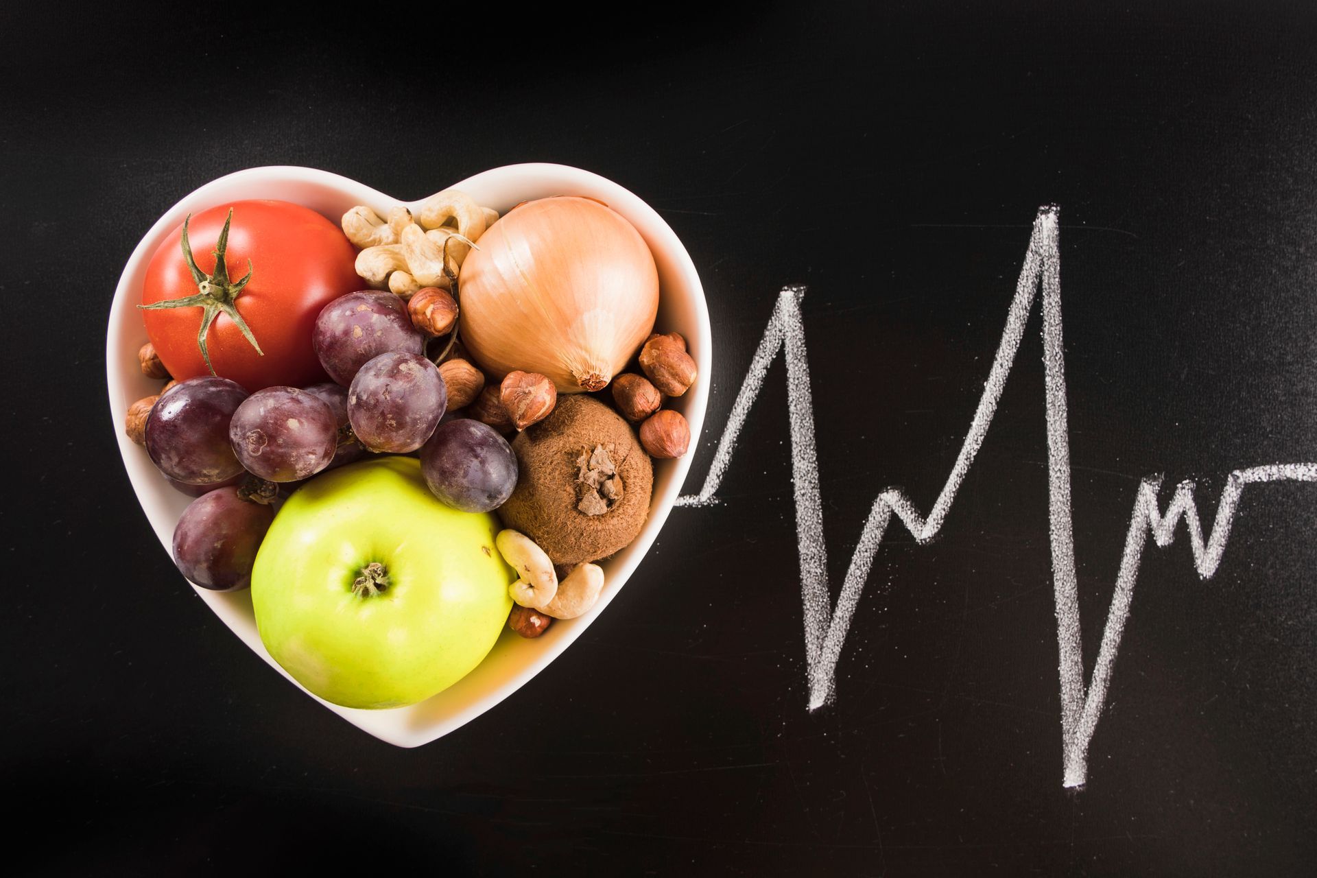 A heart shaped bowl filled with fruits and vegetables next to a heartbeat line drawn on a blackboard.