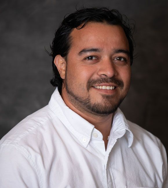 Studio headshot of a smiling man in a white button-down shirt against a dark background