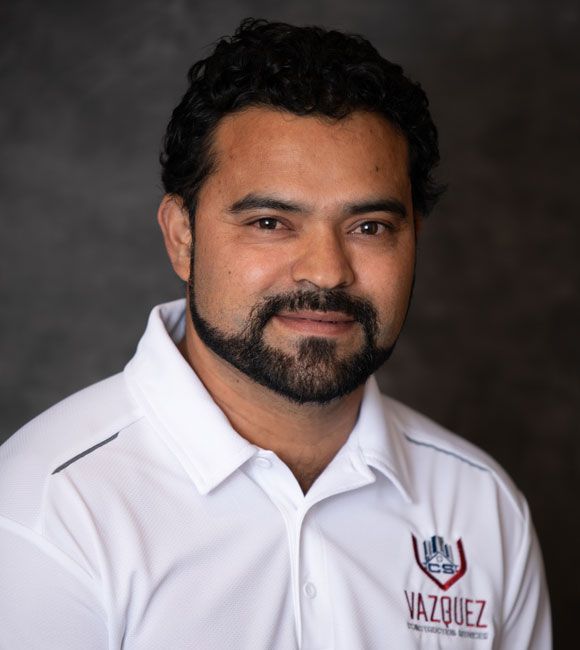 Portrait of a man in a white collared shirt with a Vazquez logo, smiling against a dark background