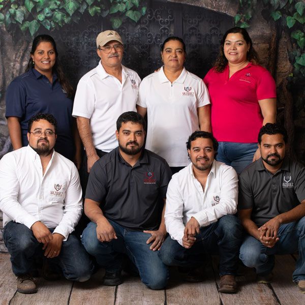 Seven coworkers pose outdoors, seated and standing in front of a leafy wall.