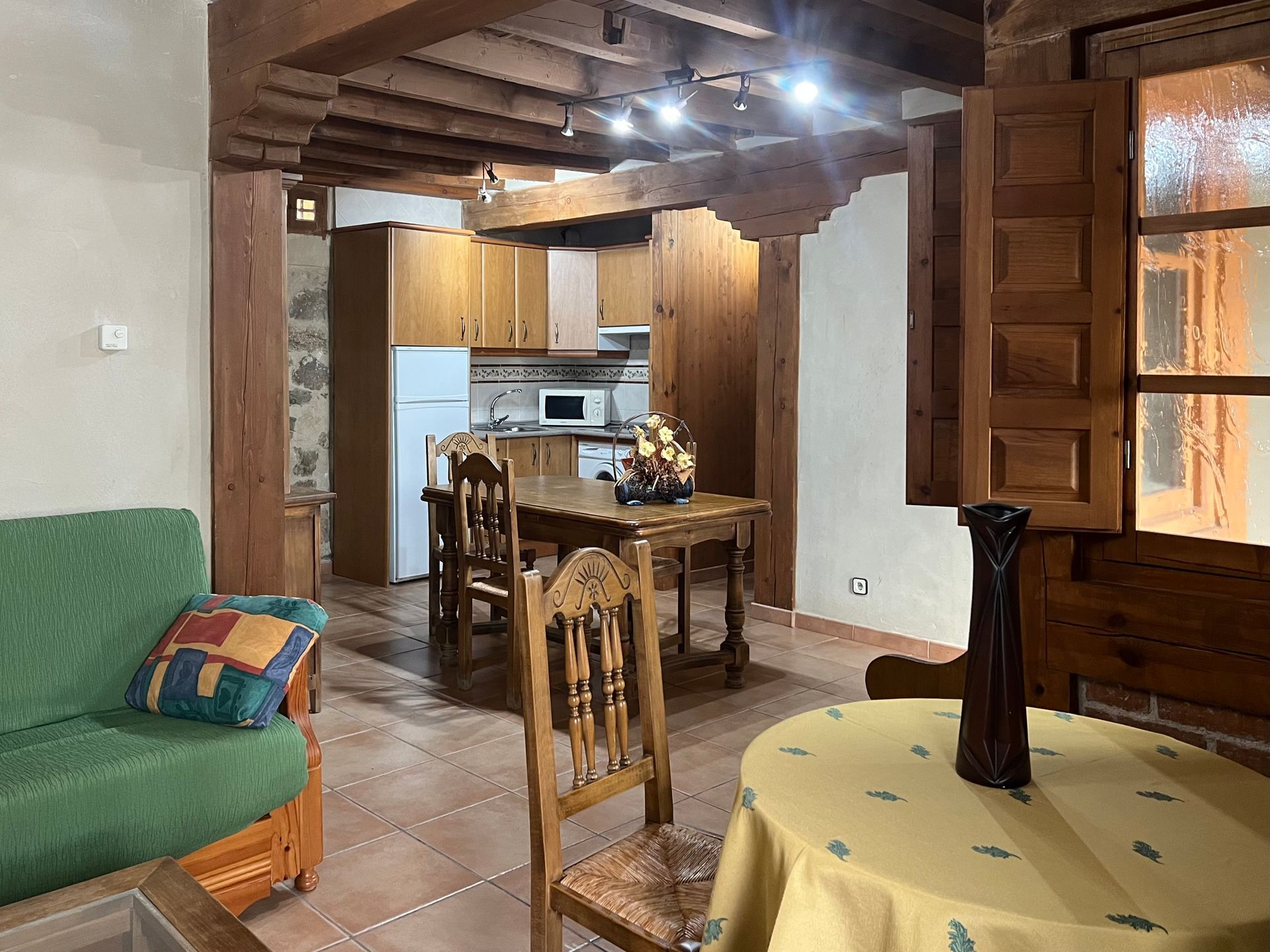 Rustic kitchen and dining area with wooden beams, tiled floor, green sofa, and table by a window.