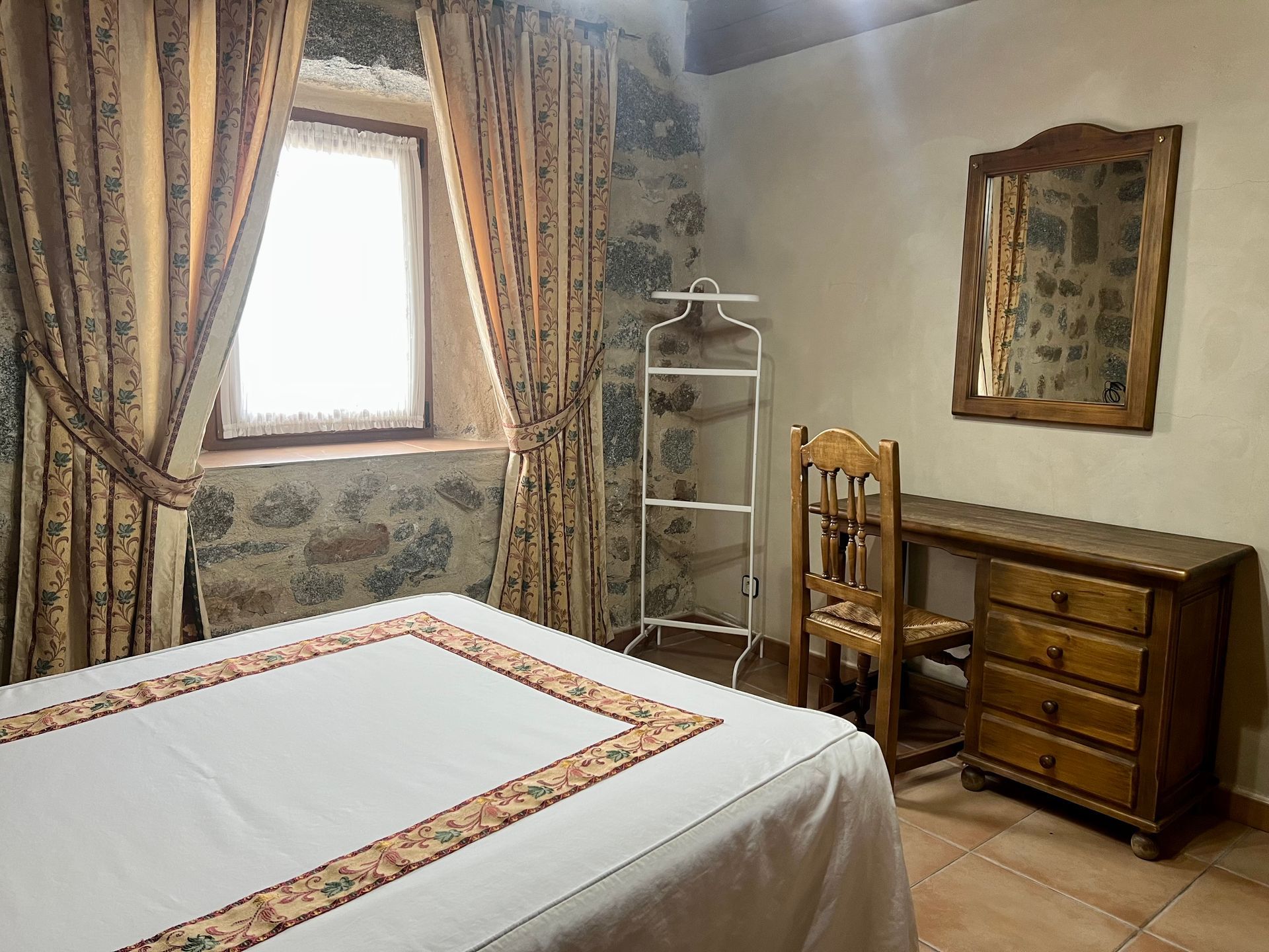 Cozy bedroom with a bed, patterned curtains, stone walls, and a wooden dresser beside a window.